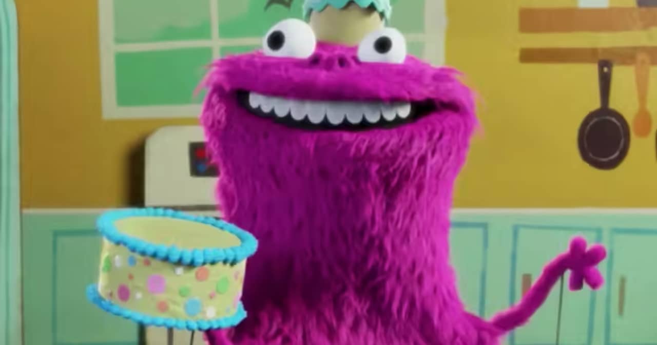 Kakie, a pink furry monster puppet with a cake hat, smiles and holds a birthday cake in a fake kitchen
